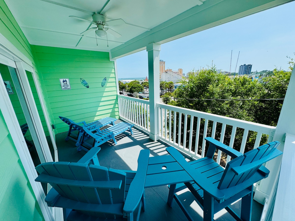A breezy balcony displays bright blue Adirondack chairs and a matching table. Sunlight illuminates the outdoor space, accentuating the fresh green walls of the home. Partial ocean views can be seen in the distance, surrounded by greenery and buildings.