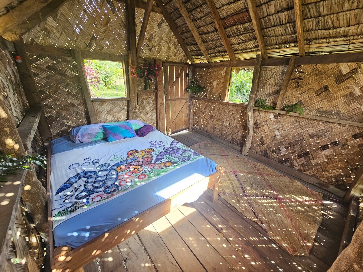 An interior view of a cozy bungalow showcases a spacious bed dressed in a colorful blanket, surrounded by woven walls of natural materials. Soft light filters through the gaps, illuminating the wooden floor. A natural mat is positioned beside the bed, enhancing the inviting space.