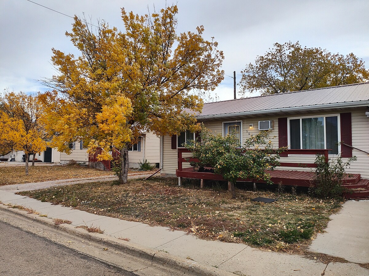 The exterior of a single-story house features a welcoming porch with a ramp, surrounded by autumn-colored trees. A paved path leads to the entrance, while a grassy area adds to the home's appeal. Nearby buildings are visible in the background.
