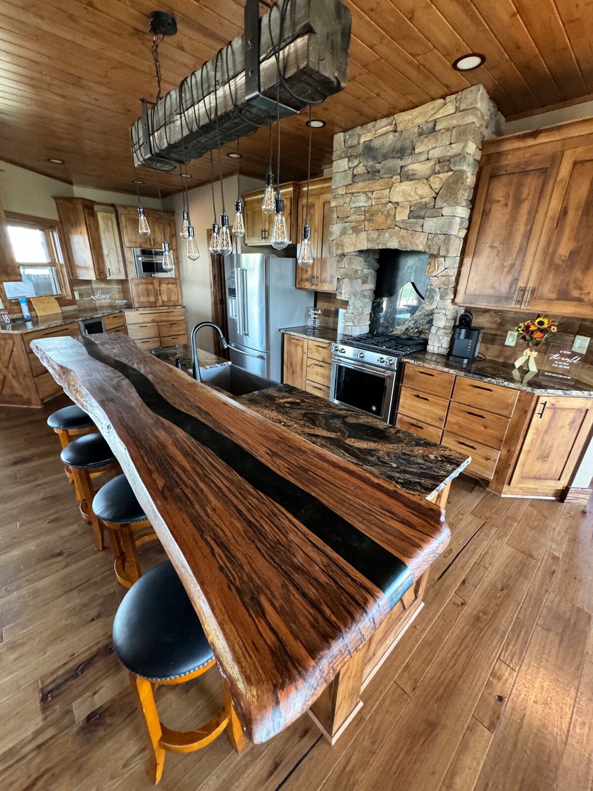 A spacious kitchen showcases natural wood cabinetry and modern stainless steel appliances. A unique live-edge wood countertop extends with a dark river inlay, surrounded by several high-backed stools. A stone fireplace adds texture to the warm wooden walls and ceiling.