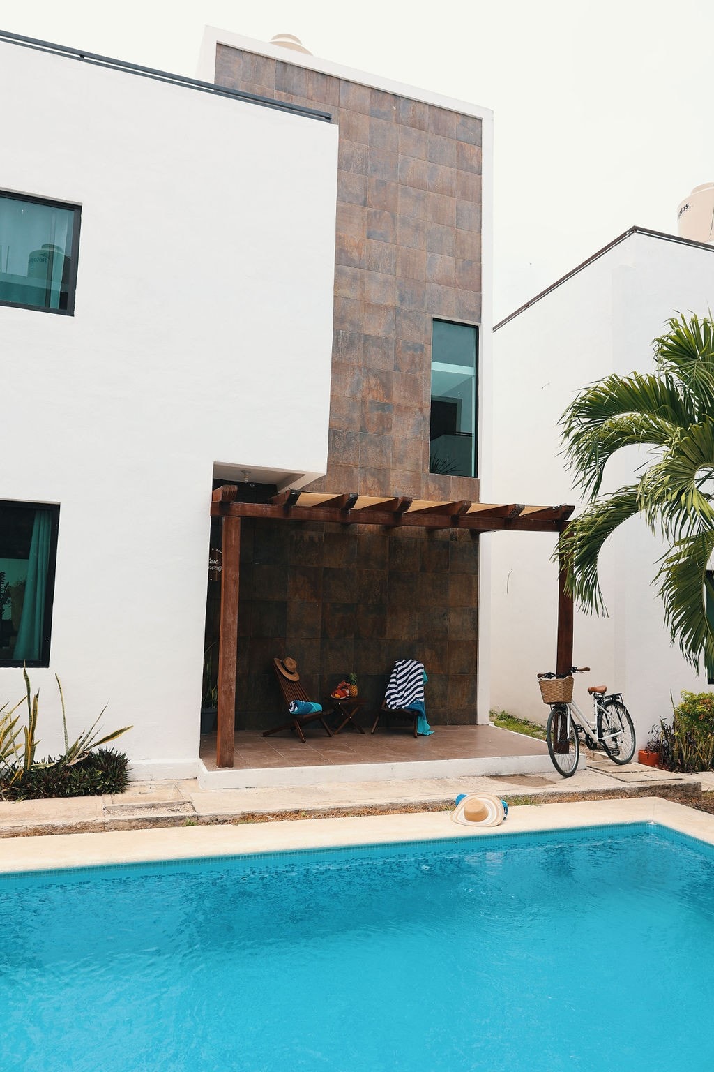 A minimalist exterior is featured, showcasing a swimming pool with clear blue water. A shaded patio area with two lounge chairs is visible beside the pool. Lush green plants and a bicycle are present, contributing to a relaxed outdoor environment.