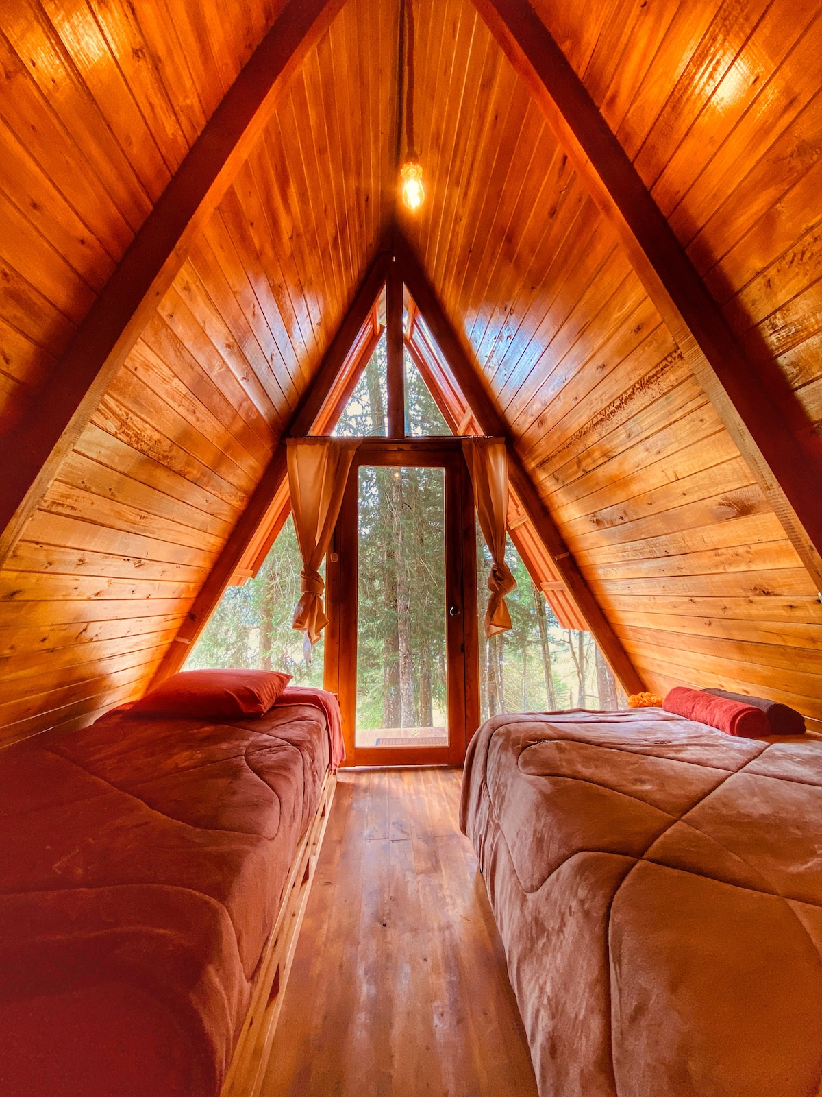A cozy sleeping area is framed by triangular wooden walls, featuring two spacious beds with soft blankets. Natural light illuminates the interior, streaming through a glass door that opens to a view of the surrounding trees.