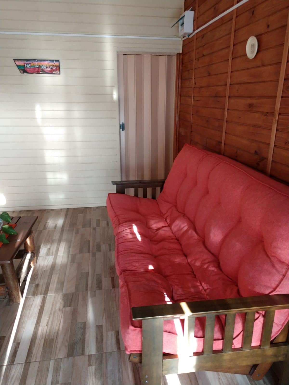 A cozy seating area features a large red couch with wooden armrests, positioned against a wooden wall. Natural light filters through a nearby door, creating a warm ambience. A coffee table sits in front, and a small plant adds a touch of greenery.