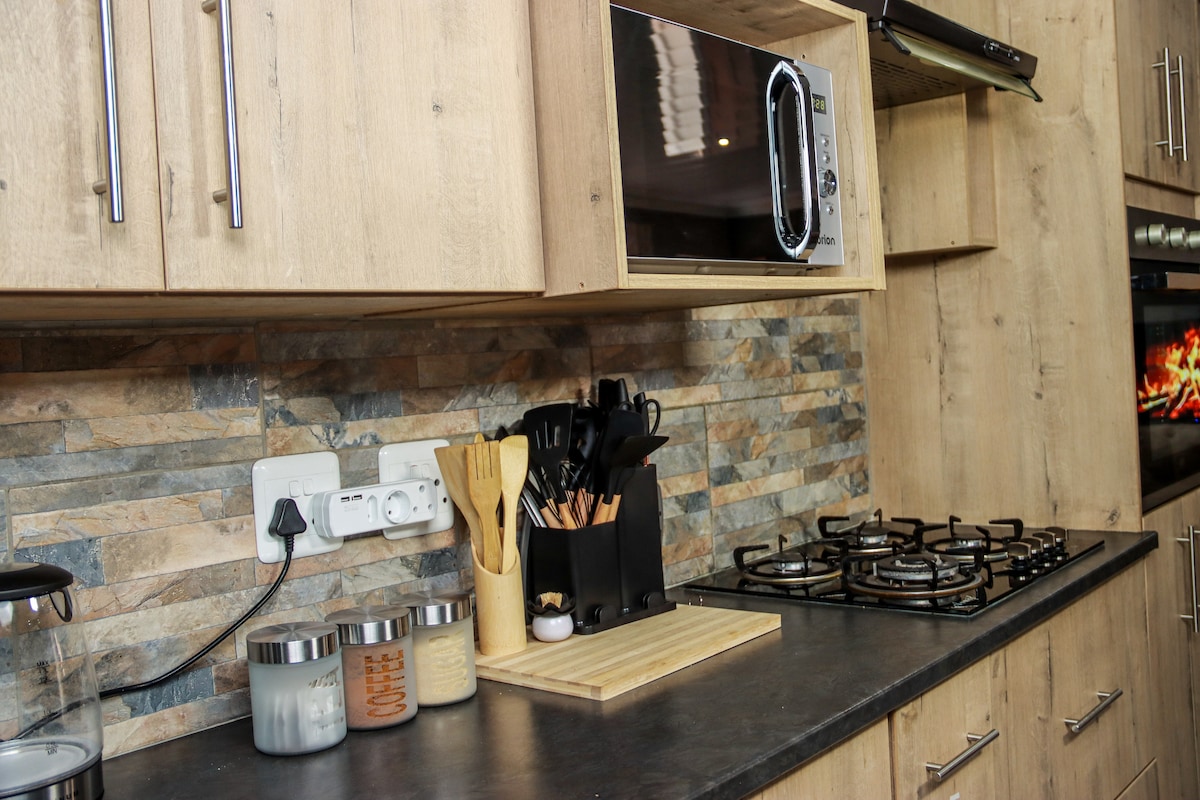 A modern kitchen is showcased with wooden cabinetry, featuring a gas stove and an electric oven. Cooking utensils and containers are neatly organized on a wooden cutting board. A countertop blender and decorative jars add functionality while creating a cohesive look.