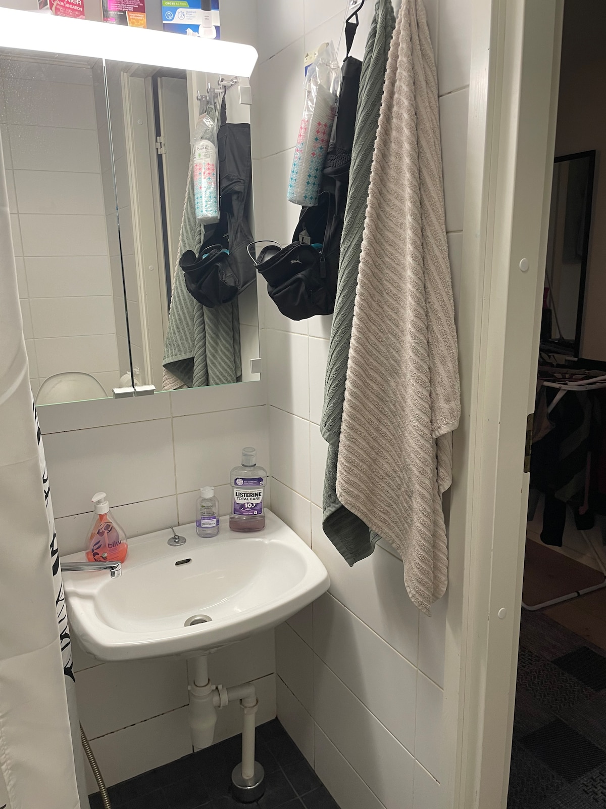 A compact bathroom is shown, featuring a small sink with toiletries placed nearby. A mirror reflects the space, while towels are hanging on hooks. The bathroom has a shower curtain partially visible, and the floor is designed with dark tiles.