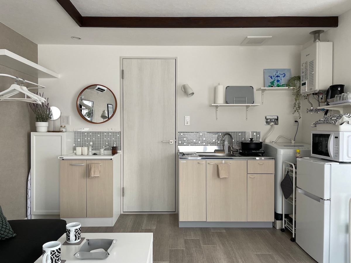 A compact kitchen area features light wood cabinetry and silver tile accents. Appliances, including a microwave and sink, are neatly organized. A round mirror reflects natural light, while a small dining table is positioned alongside a sofa in the living space.