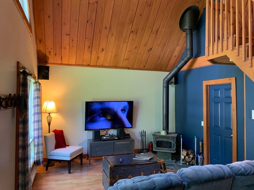 A warm living area features a high wood-paneled ceiling and a cozy sofa. A television is mounted on the wall, displaying a close-up feature. A small table with a lamp and a chair occupy one corner, while a wood stove is positioned nearby.
