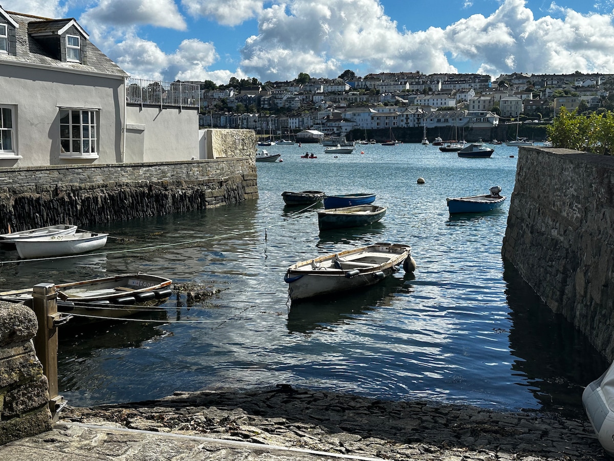 A serene view of the water, where several boats are gently bobbing in the harbor. The calm waves reflect the bright sky dotted with clouds, while quaint houses line the shoreline, providing a glimpse of the charming coastal scenery.