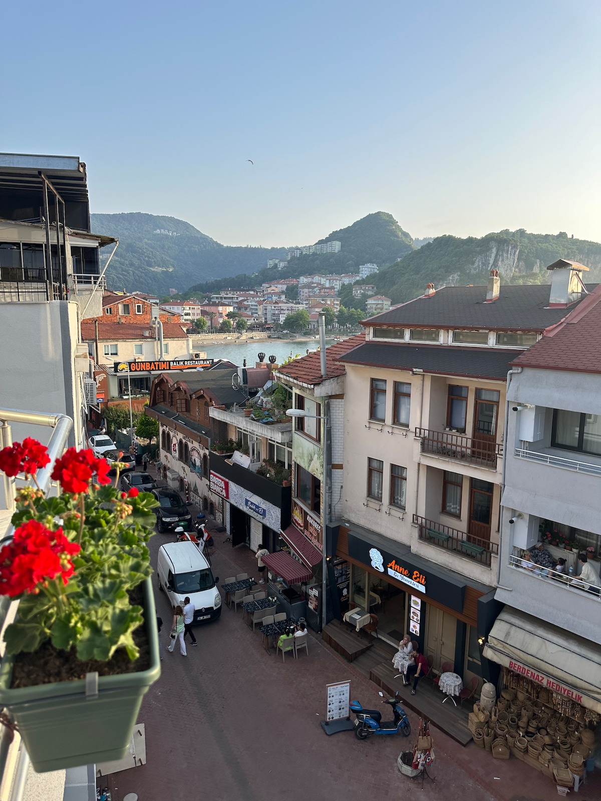 A vibrant street scene features a view of shops and buildings nestled among rolling hills. Flower pots with red geraniums are visible in the foreground, enhancing the urban landscape. The street below is bustling with activity and framed by quaint architecture.