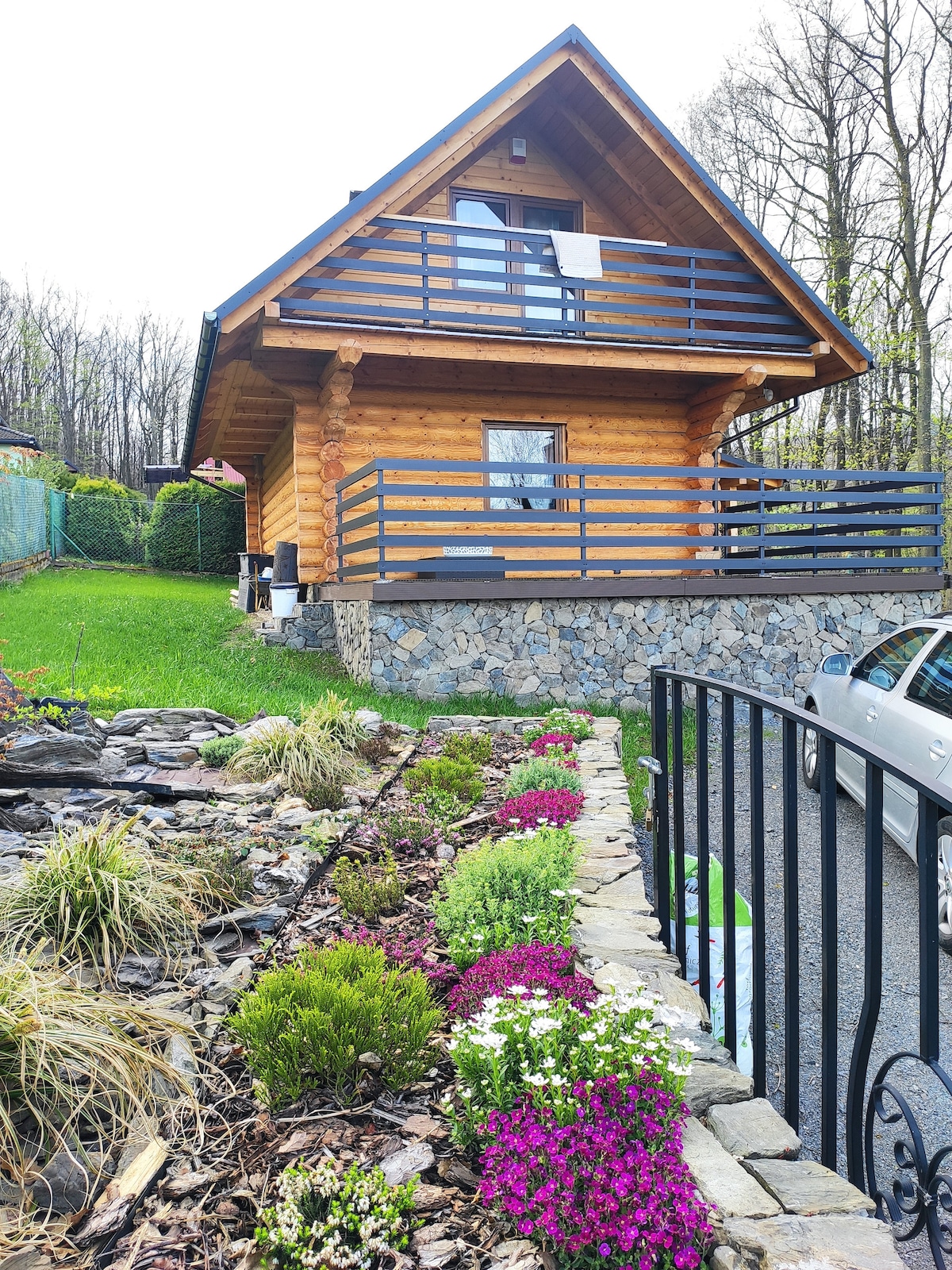 A wooden cabin with a sloped roof is showcased, featuring a stone foundation and a balcony wrapped in a blue railing. Flower beds filled with colorful blooms enhance the landscaped garden, while a paved area is visible in the foreground, indicating parking space beside the vibrant greenery.