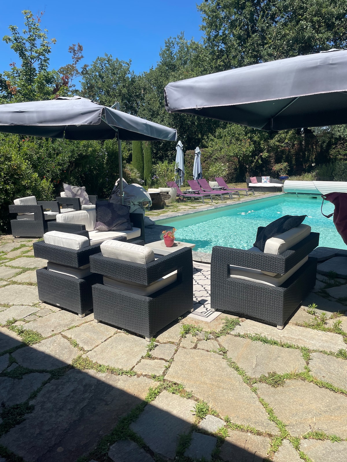 A shaded seating area is arranged with dark wicker chairs and light cushions, positioned around a stone patio. Umbrellas provide protection from the sun, overlooking a sparkling pool surrounded by greenery and sun loungers in a tranquil outdoor space.