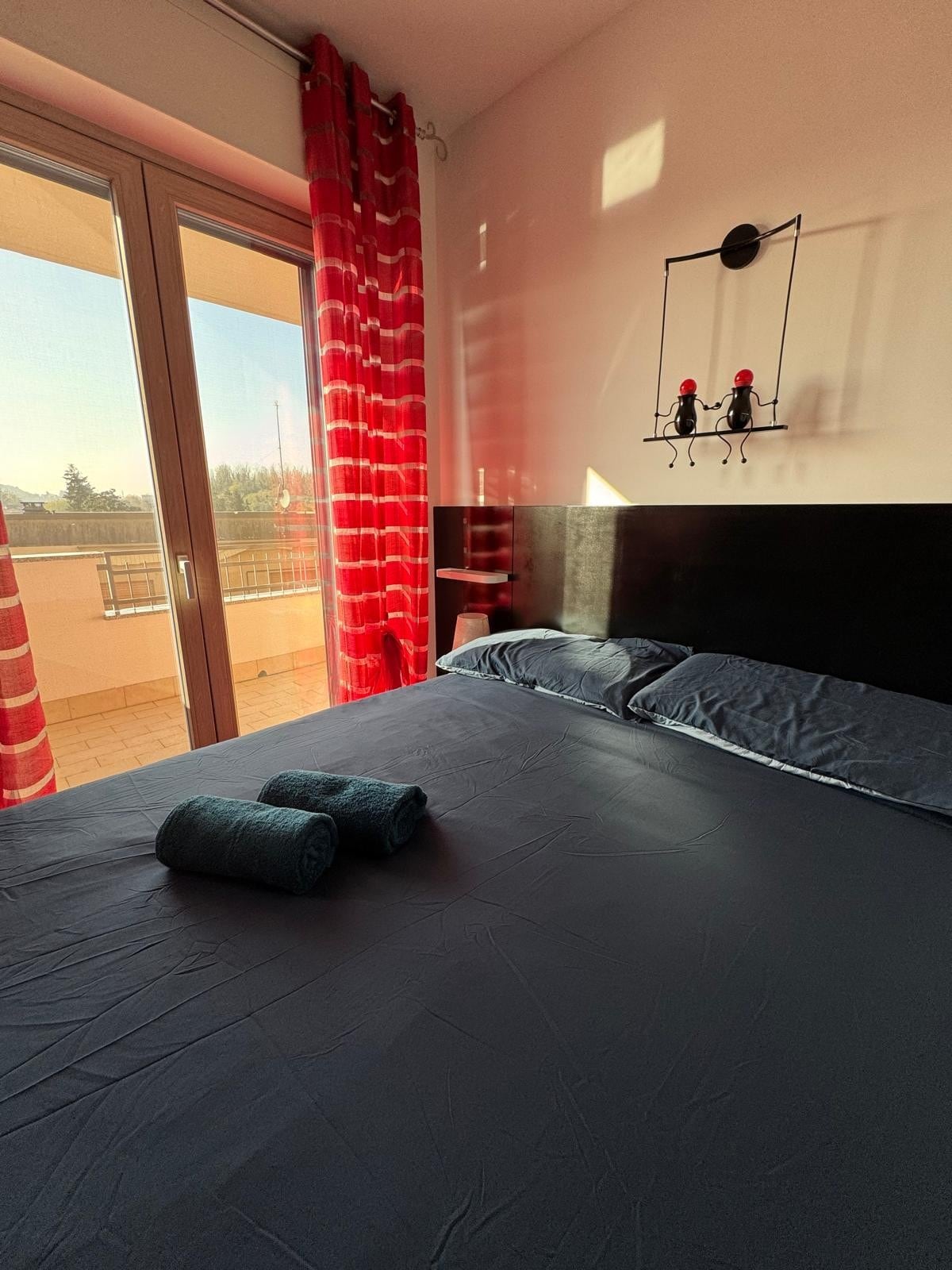A spacious bedroom is showcased, featuring a large bed covered with dark linens. Two rolled towels are neatly placed on the bed. Floor-to-ceiling glass doors provide access to a balcony, framed by vibrant red curtains that allow natural light to fill the room.