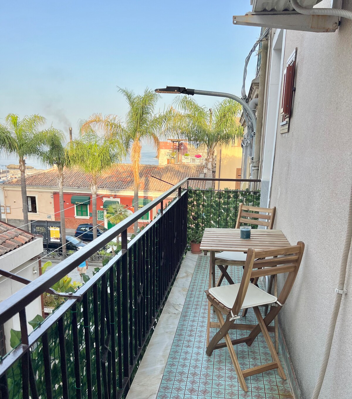 A private balcony features two wooden chairs and a small table, offering a view of palm trees and the surroundings. The tiled floor adds a decorative touch, while the railing ensures safety and openness to the outdoors.