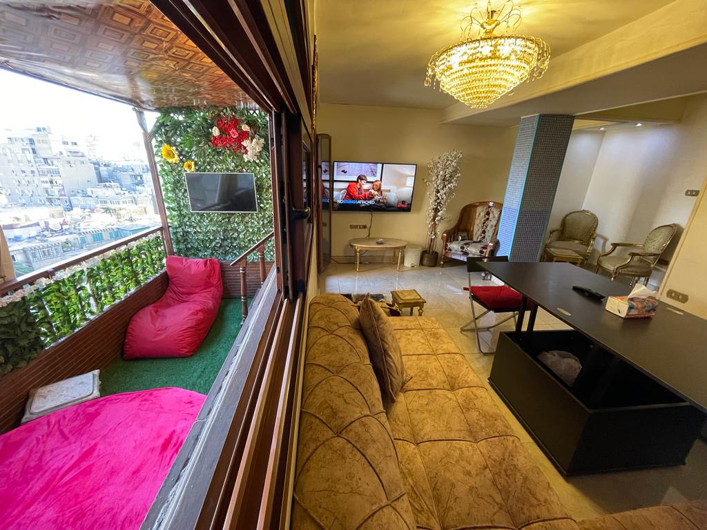 A bright and spacious living area is shown, featuring a large television mounted on the wall. Seating includes a comfortable sofa and a stylish table with chairs. A balcony with greenery and colorful bean bags enhances the connection to the outdoors.