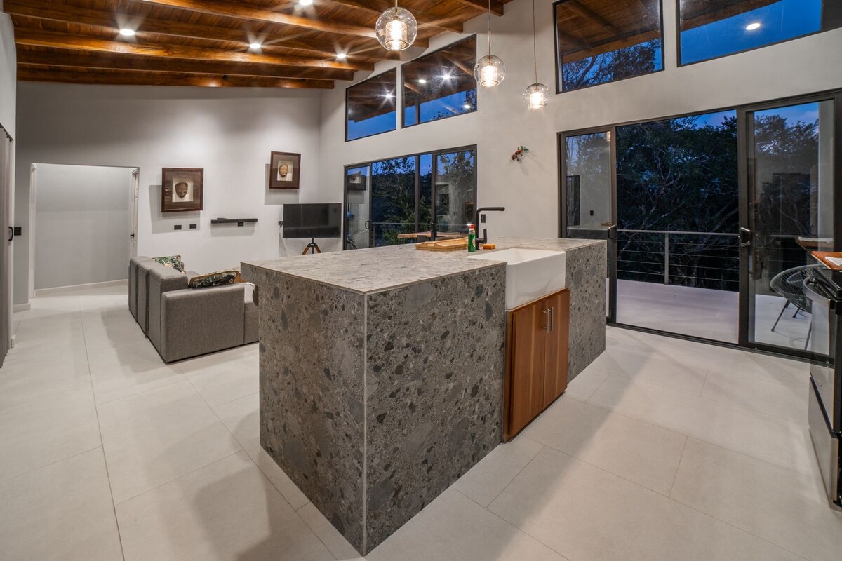 An open-plan living area features a sleek kitchen island with a textured stone surface and wooden cabinetry. Soft lighting highlights the modern decor, while large windows provide natural light and views of the outdoor space. Comfortable seating is arranged near the kitchen.