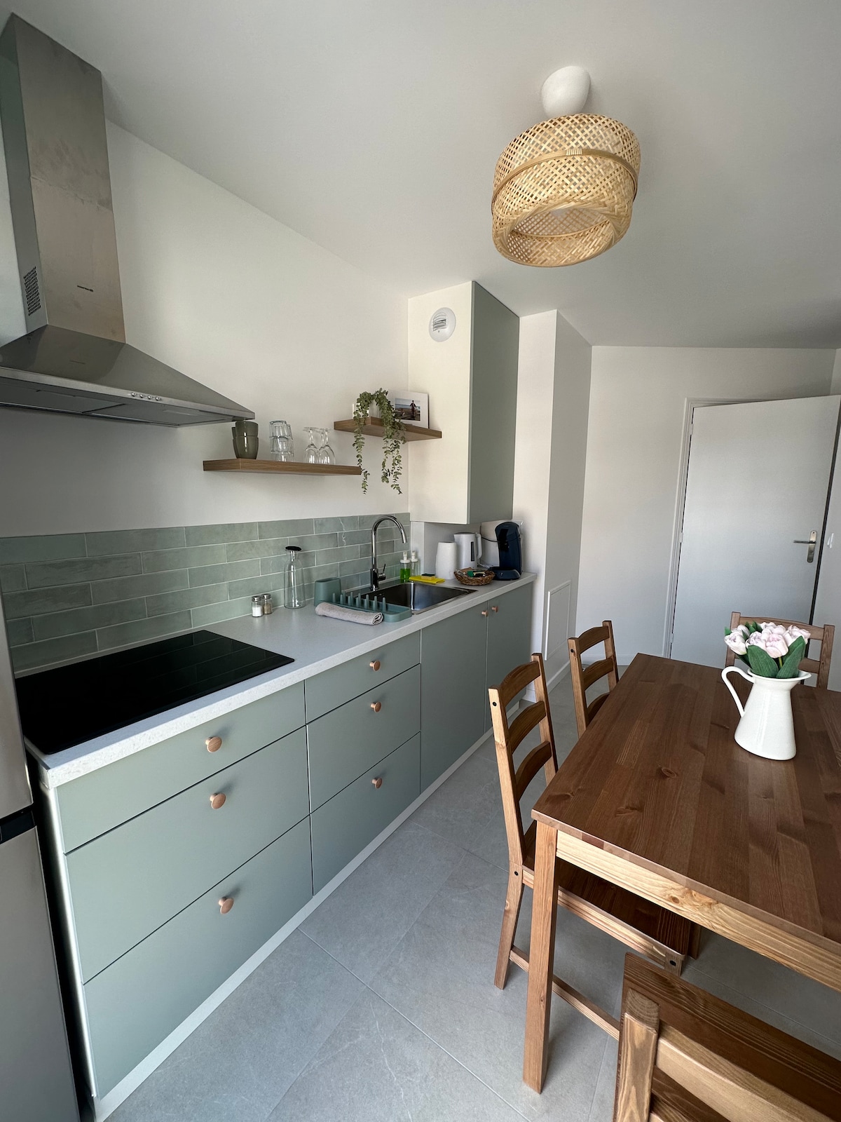 A modern kitchen is visible, featuring a minimalist design with light green cabinetry and a stylish countertop. A wooden dining table for four is set, complemented by a floral centerpiece. Appliances include a stove and kettle, with open shelving displaying glassware.