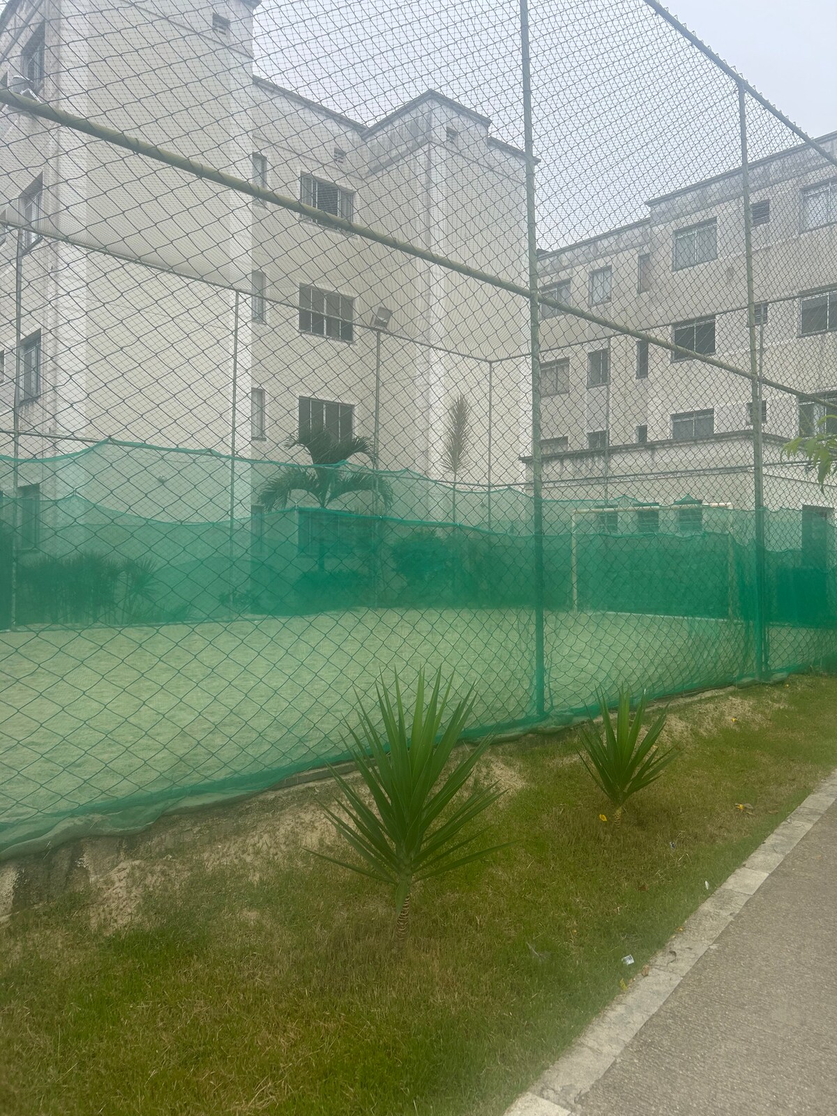 A fenced outdoor sports court is visible, surrounded by green mesh netting. The buildings behind feature multiple windows, with low landscaping including small palm-like plants along the perimeter. The sky appears overcast, providing a muted backdrop.