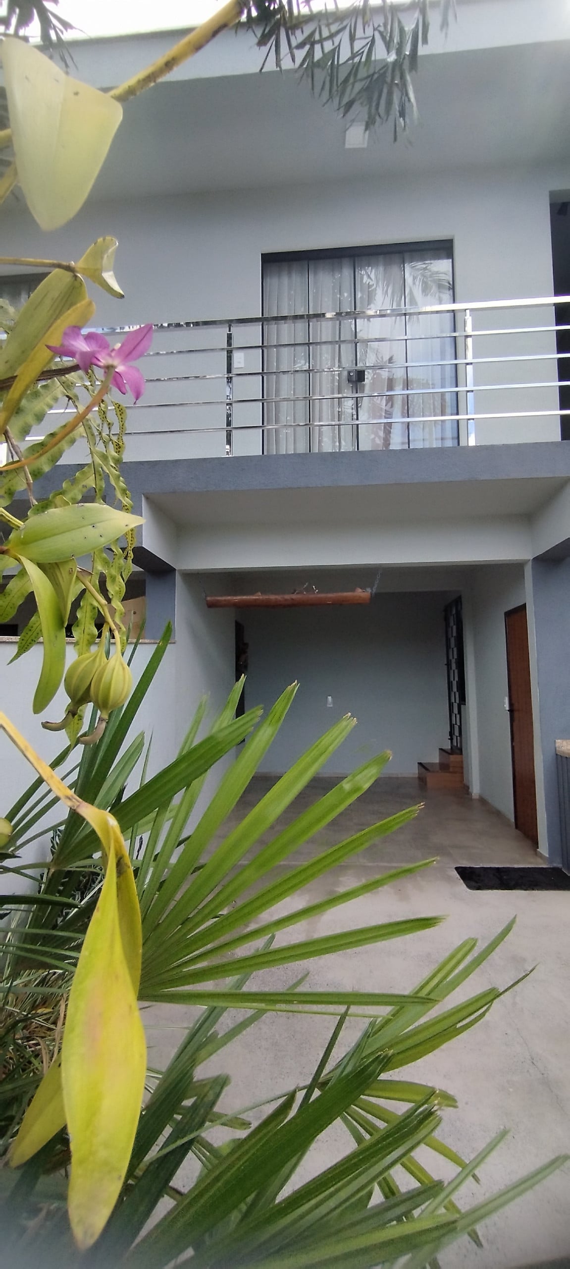 A peaceful entryway is framed by lush greenery, featuring a vertical garden with blooming orchids. The structure's modern design includes a glass door and a covered porch. Stairs lead to the upper level, offering a welcoming atmosphere for guests.