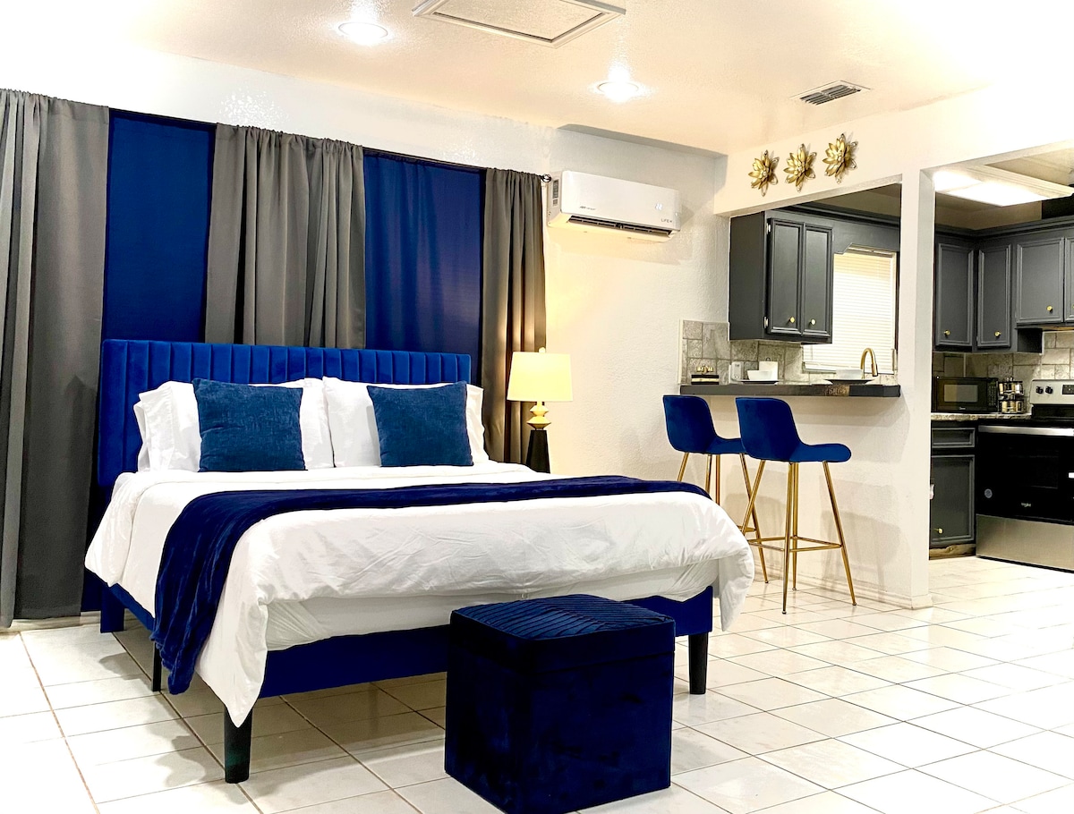 A spacious sleeping area features a plush bed with a navy blue headboard and white linens. Decorative pillows in varying shades of blue are arranged atop the bed. A small ottoman sits at the foot of the bed, and a kitchenette is visible in the background with dark cabinetry.