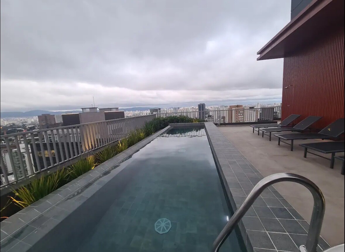 A rooftop infinity pool extends along the edge of a building, reflecting the sky and cityscape. Several lounge chairs are positioned beside the pool with greenery peeking through. A railing provides a clear view of the sprawling urban landscape in the background.