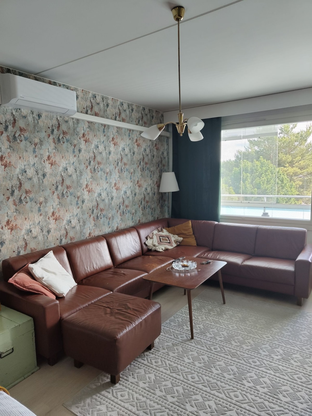 A cozy living room is featured with a brown leather sectional sofa and a low wooden coffee table. A patterned rug covers the floor, while a large window offers natural light. Decorative pillows and a small clear glass bowl are placed on the table, complemented by a wall adorned with floral wallpaper.