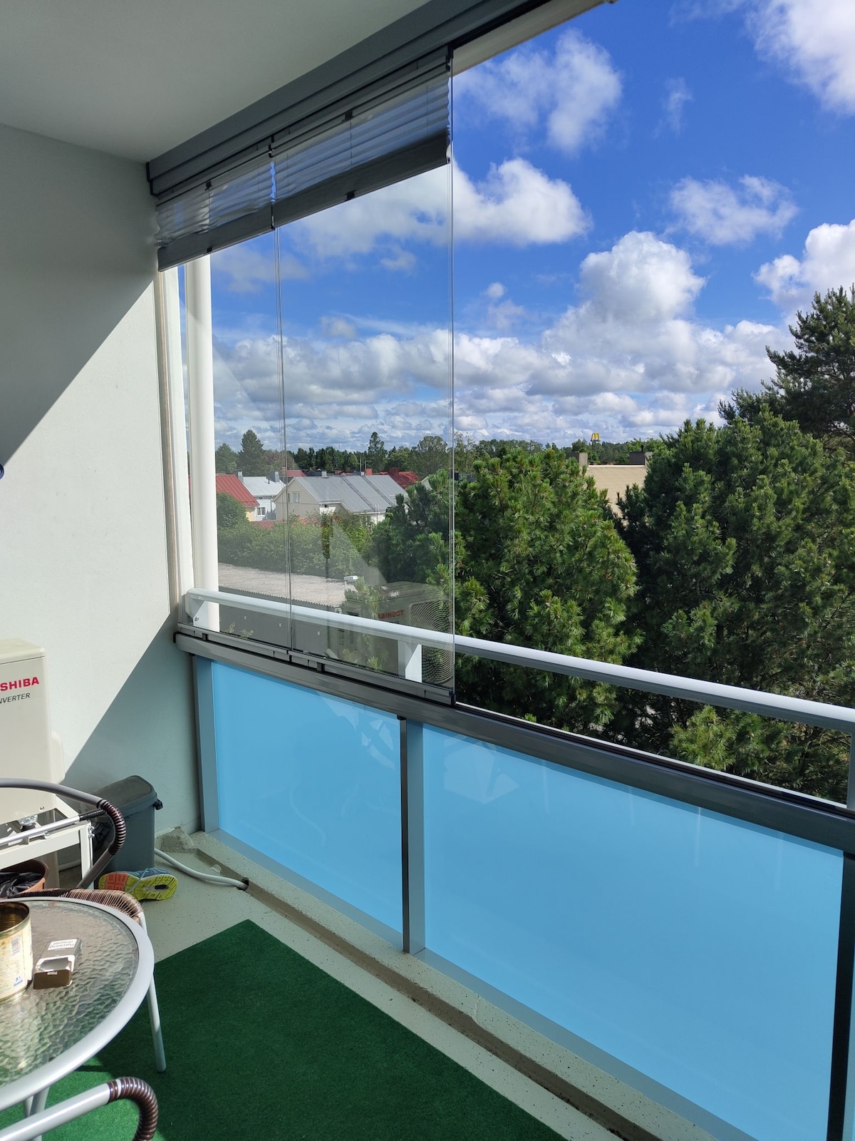 A balcony area is shown with glass sliding panels allowing for unobstructed views of the sky and surrounding landscape. A small round table and a few chairs are present on a green carpeted floor, creating a space for relaxation.