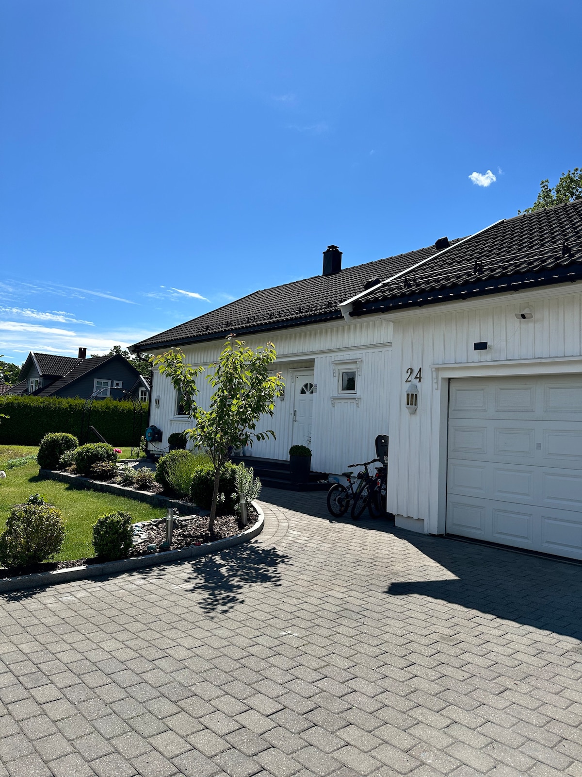 The exterior of a well-maintained house is displayed, featuring a light-colored façade and a driveway lined with neat hedges. A manicured lawn with bushes enhances the inviting appearance, and a garage door is visible on the right side. Two bicycles are parked near the entrance.