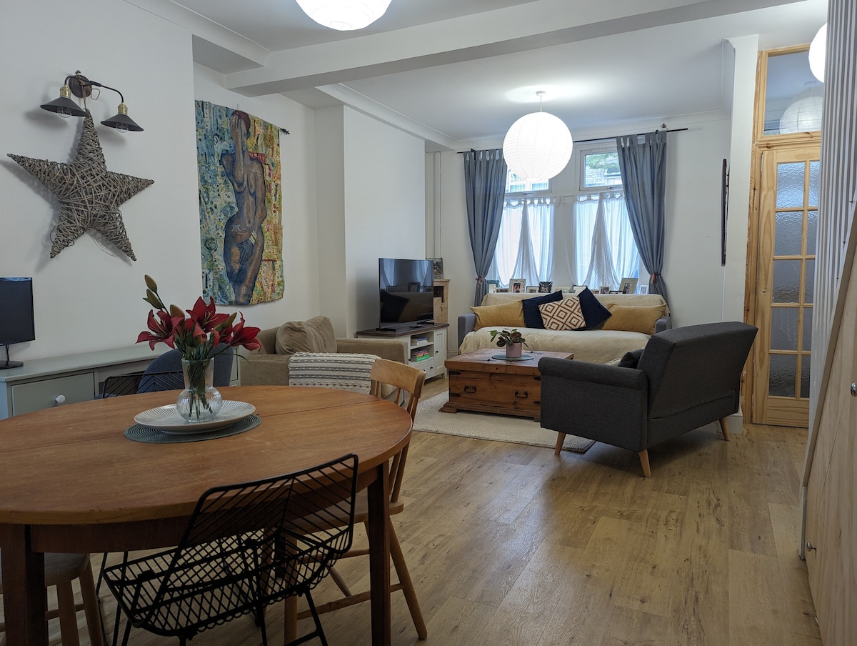 An open plan living area is shown, featuring a round dining table set for four, a cozy seating arrangement with a beige sofa and grey chairs. Natural light streams through large windows adorned with grey curtains, while a decorative star and vibrant wall art add character.