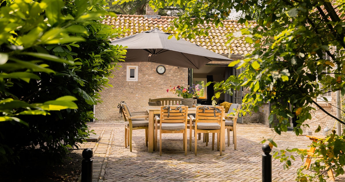 A spacious outdoor dining area is framed by lush greenery, featuring a large umbrella over a wooden table surrounded by six chairs. Brick paved flooring leads to a rustic structure in the background, which is partially obscured by surrounding foliage.