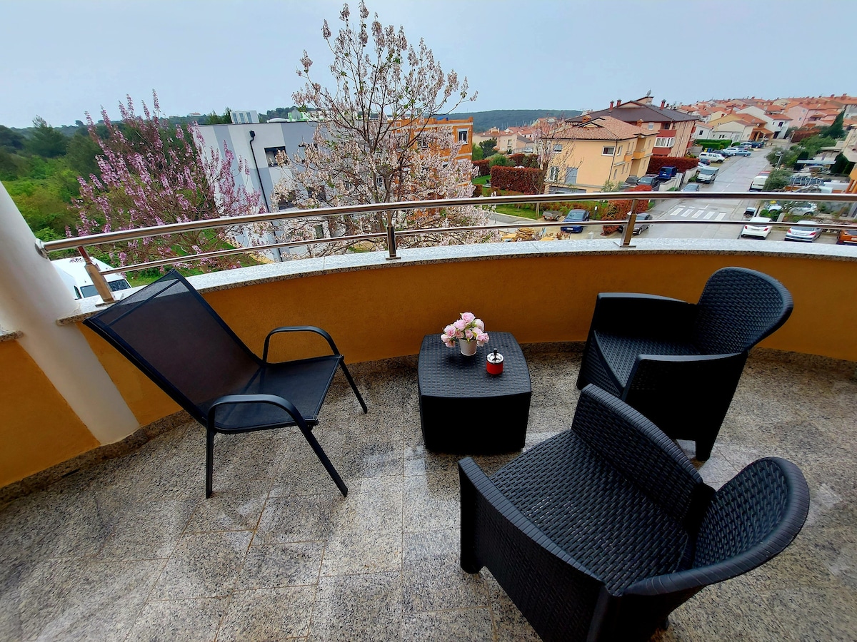 A spacious balcony is furnished with two lounge chairs and a small table, set on a smooth surface. A view of the surrounding neighborhood and flowering trees is visible beyond the balcony's railing, creating a peaceful outdoor space.