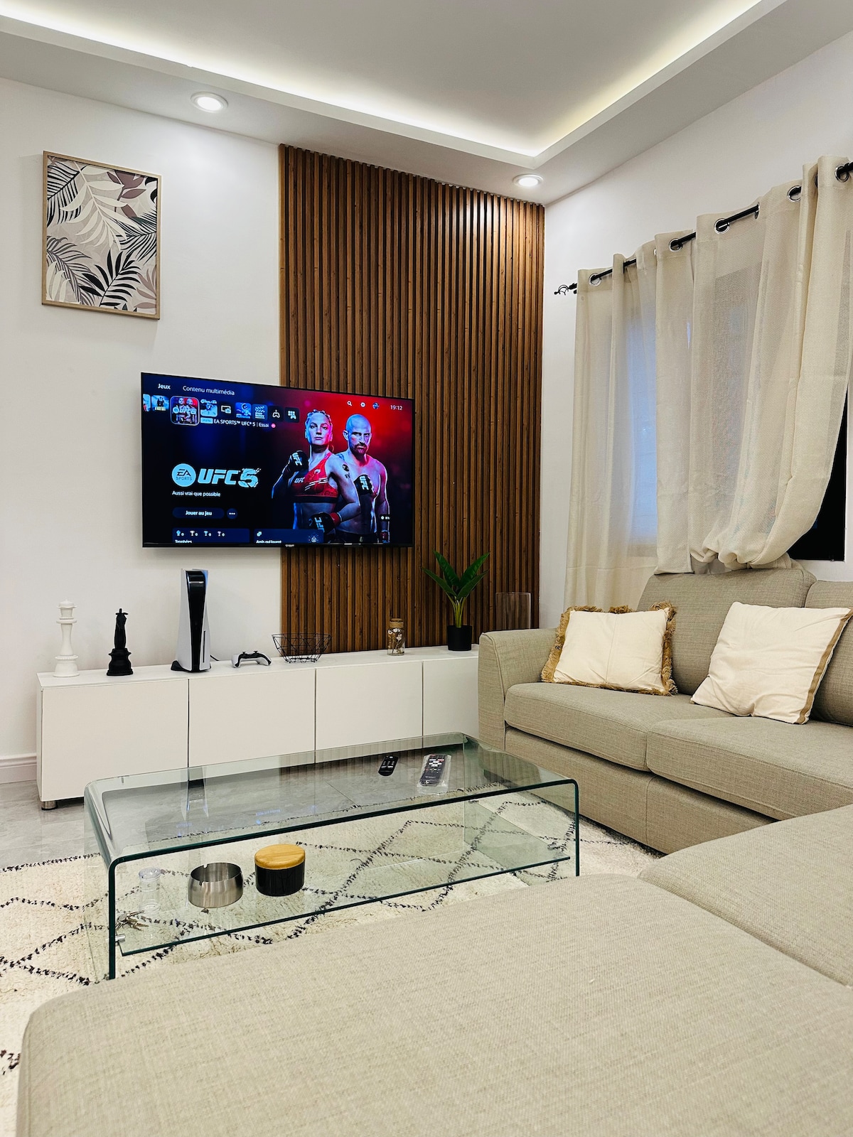 A comfortable living area is showcased, featuring a light-colored sectional sofa and a glass coffee table. A flat-screen television is mounted on a wood-paneled wall, displaying a vibrant image. Soft curtains frame the window, allowing for natural light to fill the space.