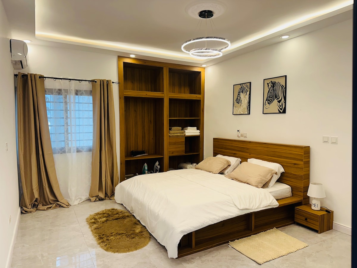 A spacious bedroom features a king-size bed with neutral bedding. Wooden shelves are positioned against the wall, displaying towels and decorative items. Natural light enters through a window with sheer curtains, while soft lighting is provided by a modern overhead fixture.
