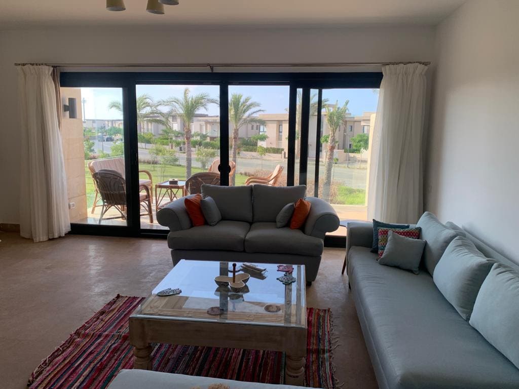 A spacious living room features a light grey sofa complemented by colorful cushions and a rustic coffee table. Large glass doors lead to a furnished balcony with seating and views of palm trees and surroundings. Natural light fills the room through the curtains.