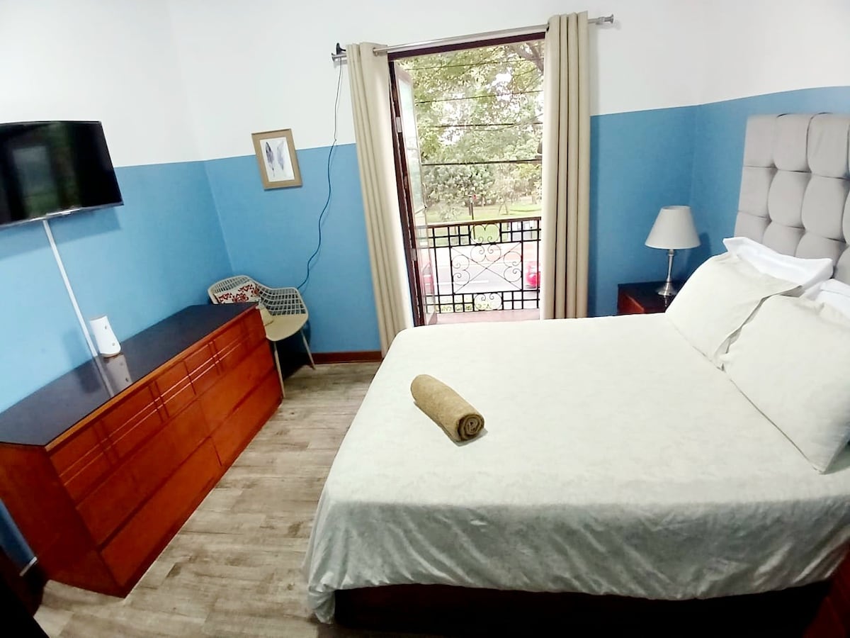 This welcoming bedroom features a spacious bed with a light-colored coverlet and a rolled towel placed on top. A television is mounted on the wall opposite the bed, and a wooden dresser stands nearby. Natural light filters through the window, surrounded by light curtains.