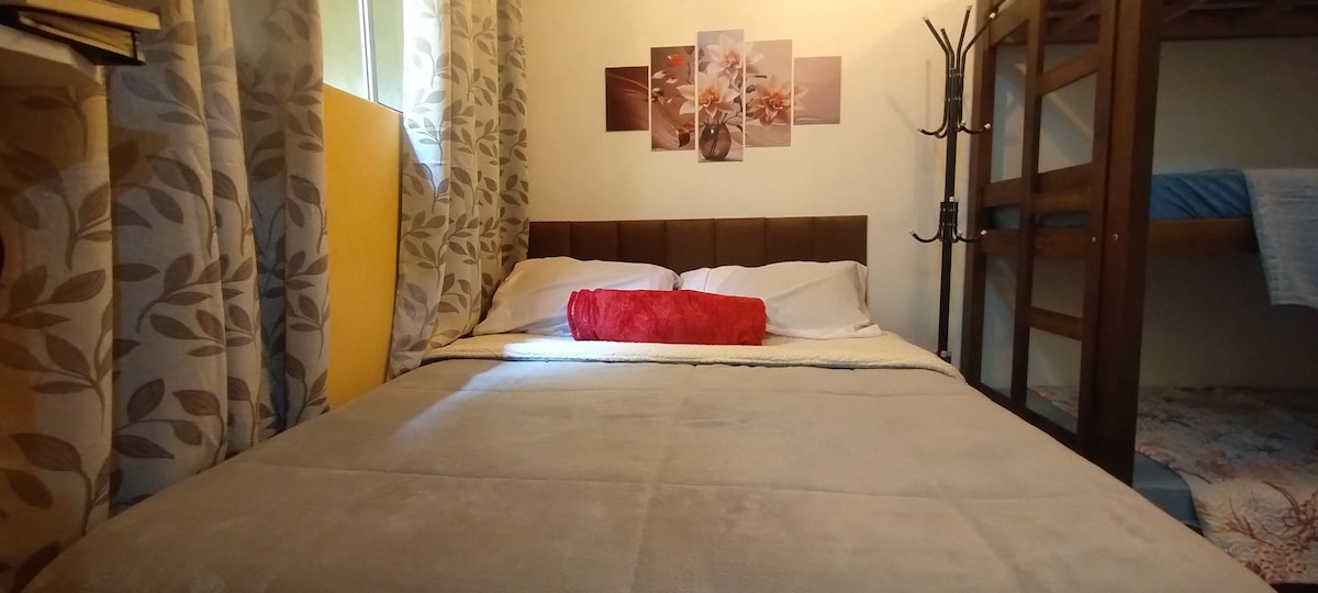 A cozy bedroom features a spacious double bed, adorned with a light blanket and a red accent pillow. Above the bed, a series of wall art provides a warm touch. A bunk bed is positioned in the corner, offering additional sleeping space.