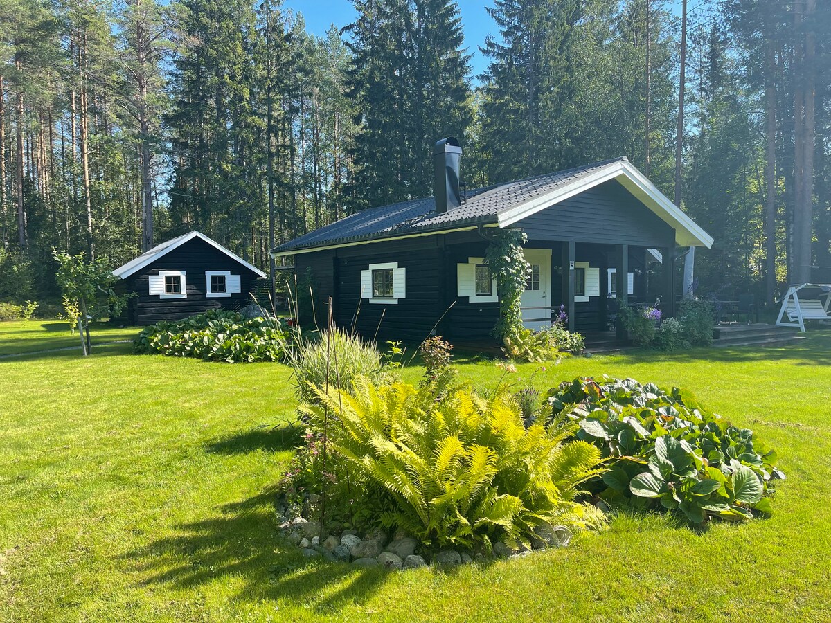 A charming black-painted house is surrounded by lush greenery and gardens. The landscape includes various plants and a manicured lawn, while the home features a welcoming porch. A second structure is visible nearby, nestled among tall trees that provide natural shade.