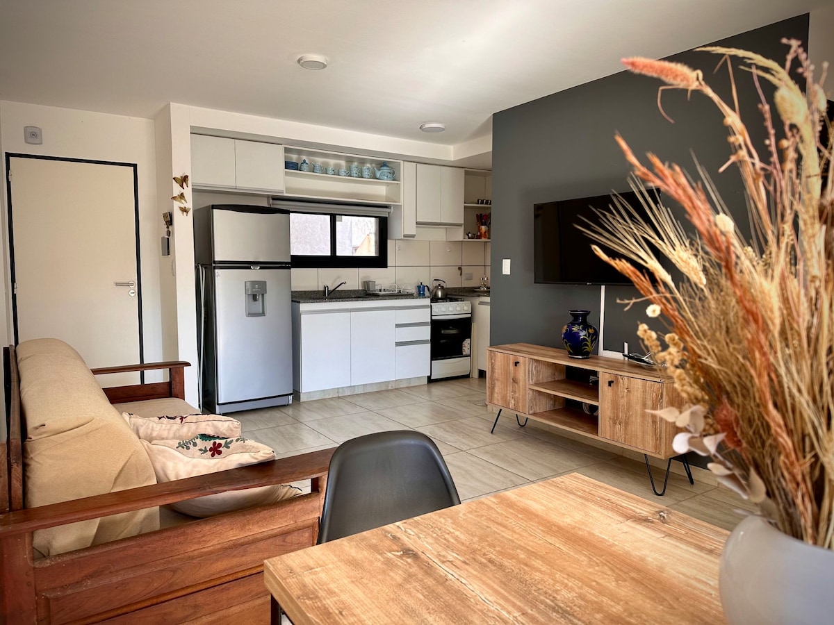The living area features a spacious couch facing a mounted television. A wooden dining table is positioned nearby, complemented by decorative dried plants. The kitchen area includes stainless steel appliances and cabinets, with natural light entering through a window above the sink.