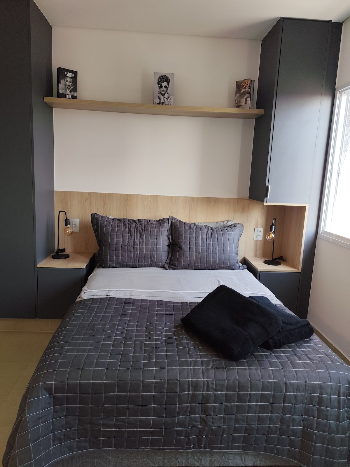 A cozy sleeping area is presented with a spacious bed adorned with gray bedding. Shelving units above the bed display decorative items. Soft lighting is provided by bedside lamps. Natural light enters through the adjacent window, enhancing the room's calm ambiance.
