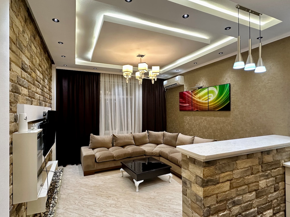 A modern living area is characterized by a spacious sectional sofa and an elegant coffee table. The room features a stone accent wall, soft lighting from recessed fixtures, and a colorful wall art piece. Natural light filters through sheer curtains, enhancing the ambiance.