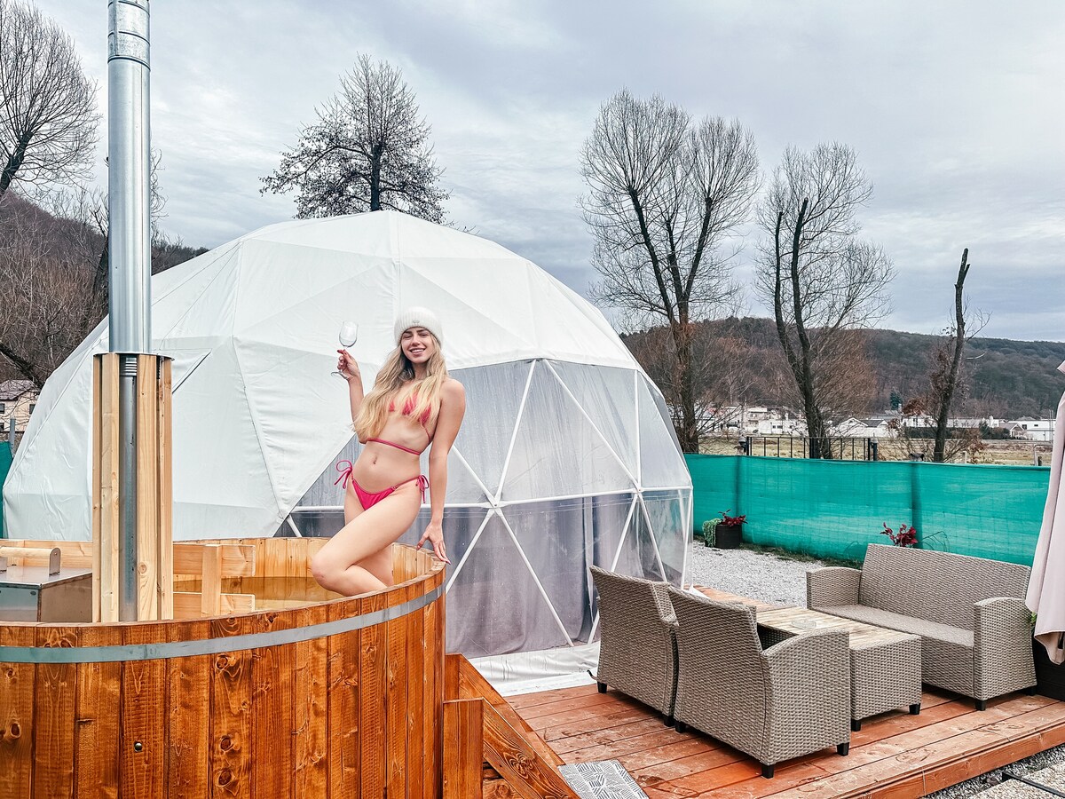 A geodesic dome stands amid natural surroundings, featuring a wooden hot tub on a wooden deck. Nearby, a seating area with comfortable, neutral-toned furniture invites relaxation. Trees line the background, enhancing the tranquil atmosphere of the outdoor setting.