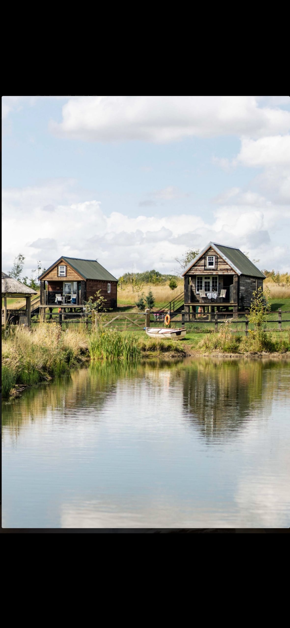 Two unique lodges are positioned by a tranquil lake, offering a serene retreat. Each lodge features large windows and a porch with seating, surrounded by lush greenery and a calm water reflection that enhances the peaceful setting.