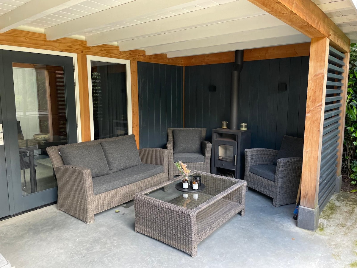 A sheltered seating area features a cozy arrangement of dark wicker furniture, including a spacious sofa and two chairs. A glass-top coffee table sits in the center, complemented by a small wood stove, offering a relaxing space to enjoy the outdoors.