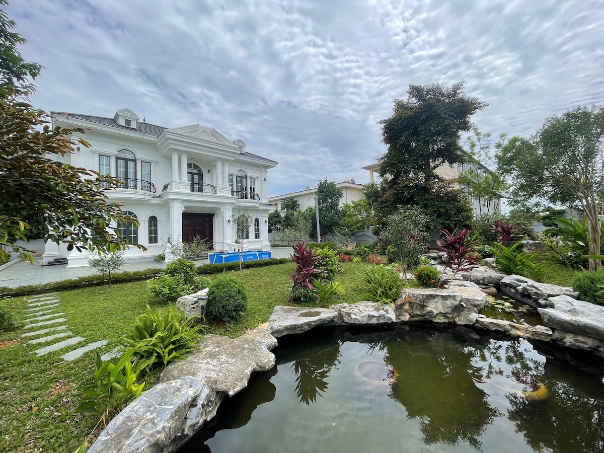 A two-story villa is set amidst a lush garden, featuring neatly arranged greenery and vibrant plants. A serene pond with visible water features is located in the foreground. The sky is overcast, adding a soft light to the tranquil setting.