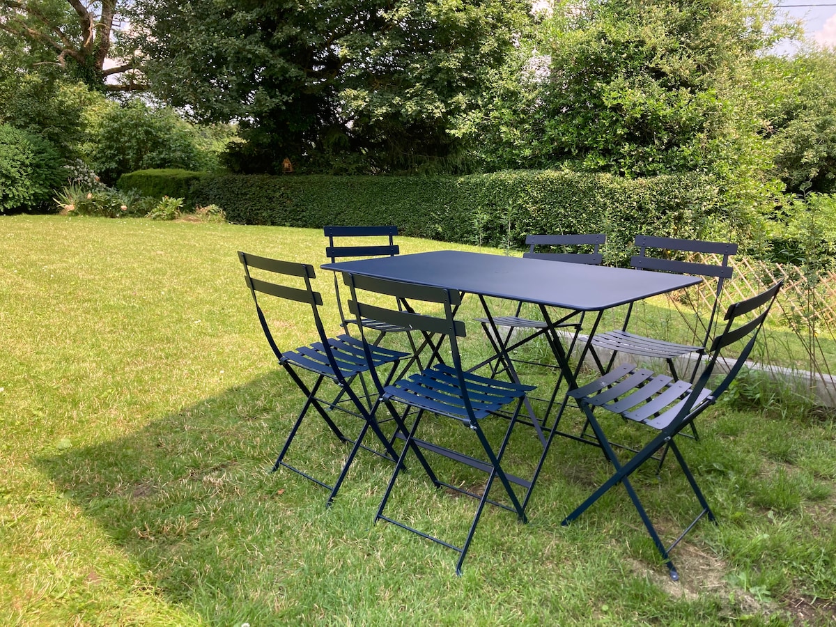 A folding table made of metal is surrounded by six matching chairs, all arranged on a grassy area. Lush greenery is visible in the background, providing a serene outdoor setting. The well-maintained lawn enhances the inviting space for relaxation or dining.