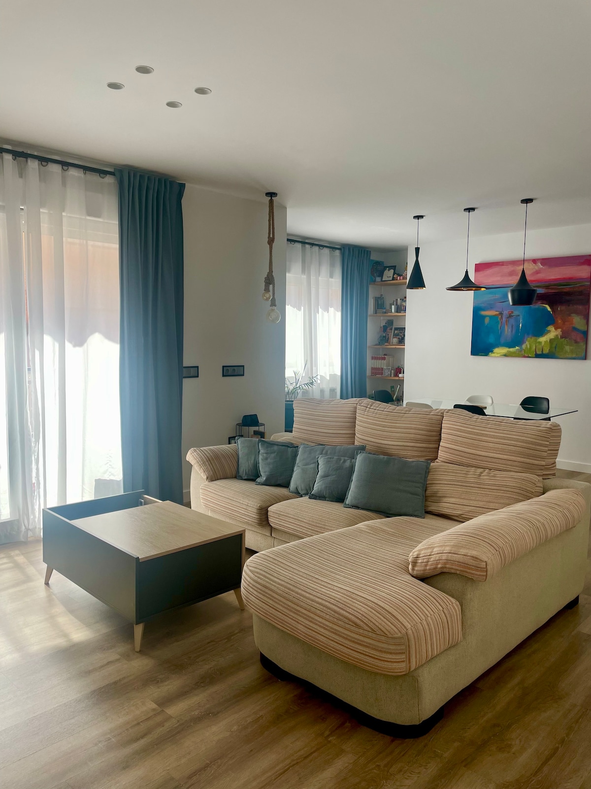 A comfortable living area features a large sectional sofa adorned with cushions. Natural light floods the space through tall windows with sheer curtains. A modern coffee table meets wooden flooring, while a dining area is visible in the background, highlighted by three pendant lights.