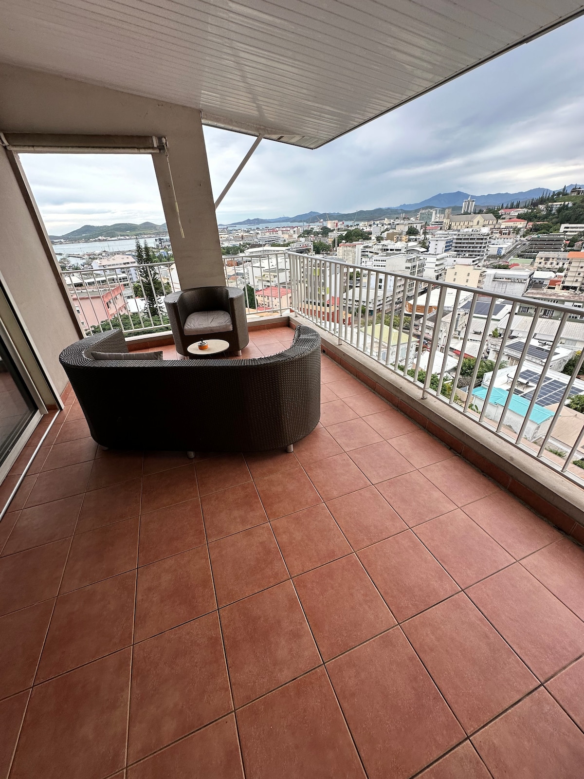 A spacious terrace is featured, equipped with comfortable seating. The surface is tiled, and a scenic view of the surrounding area and distant mountains is visible beyond the railing. The sky appears overcast, suggesting a tranquil atmosphere.