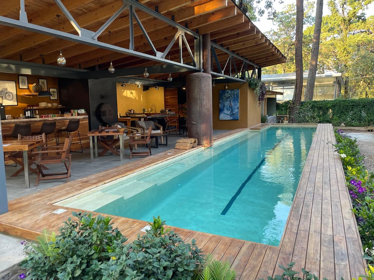 A serene outdoor area features a sleek, rectangular pool bordered by wooden decking and surrounded by lush greenery. An open-air dining space with wooden tables and chairs is visible under a high wooden roof, complemented by soft lighting. The backdrop includes a warming fireplace, framed artwork, and natural decor.