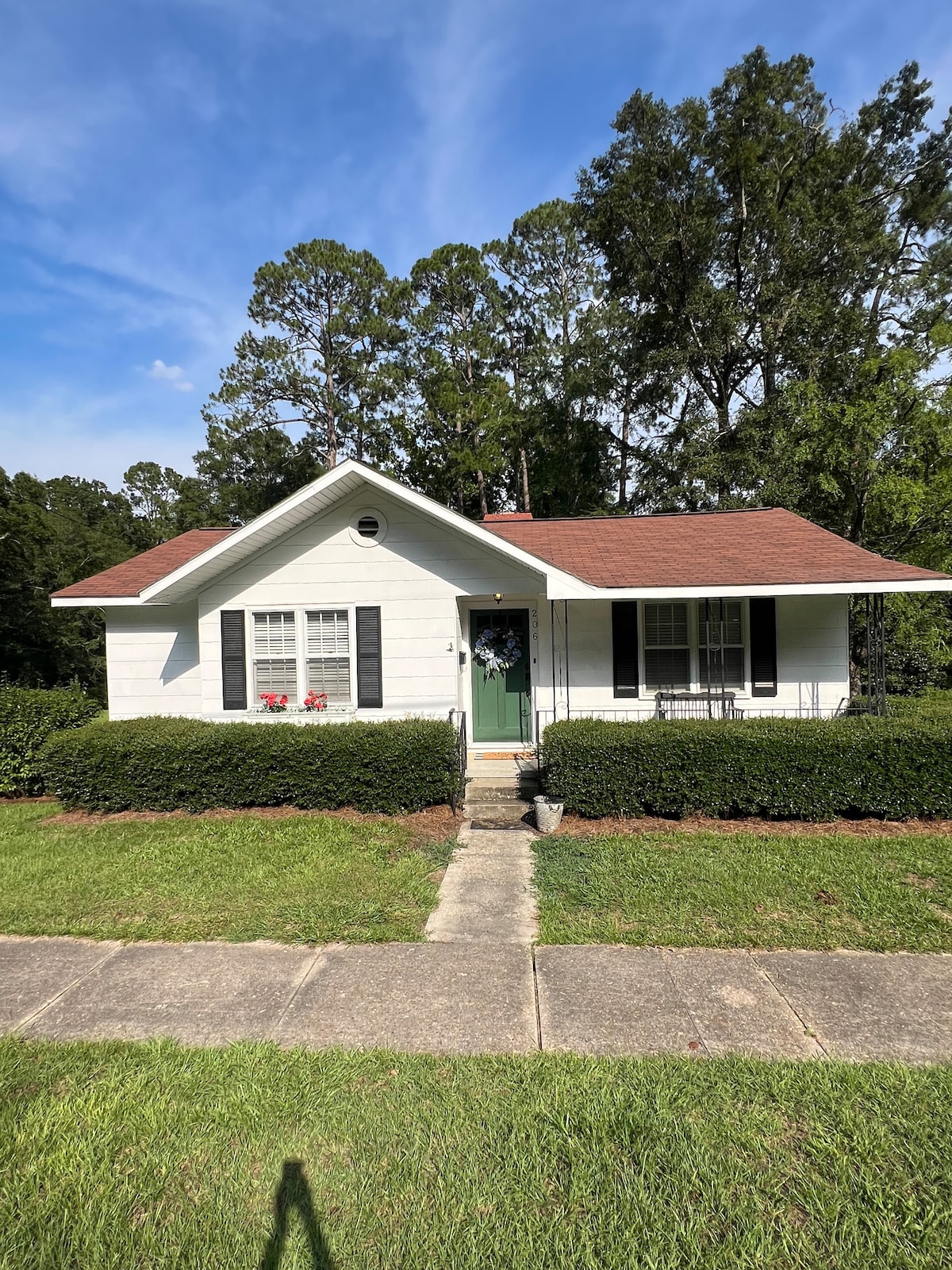 A single-story house with a white exterior is framed by neatly trimmed bushes and a well-maintained lawn. A central front door is flanked by two windows, with flower boxes added for color. The roof is covered with brown shingles, and trees provide greenery in the background.