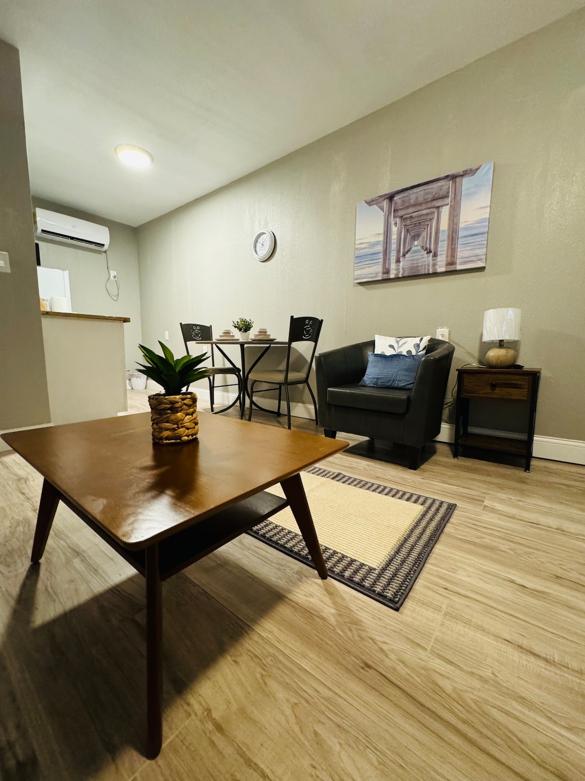 A cozy studio apartment is depicted, featuring a small dining table with two chairs and a black armchair. A wooden coffee table sits in the foreground, complemented by a decorative plant. Soft lighting enhances the neutral-colored walls, with a framed wall art piece visible in the background.