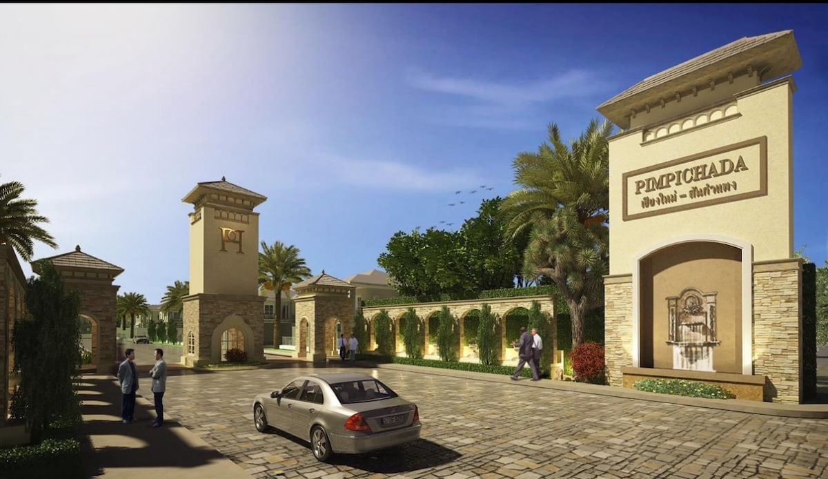 The entrance to a serene residential community is showcased, featuring stone columns and a decorative water fountain. Palm trees and well-manicured landscaping surround the area, with a parked car and individuals walking along the paved pathway, suggesting a welcoming atmosphere.